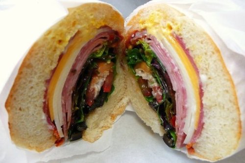 A cross-section of a sandwich filled with layers of meats, cheese, and fresh vegetables on soft bread.