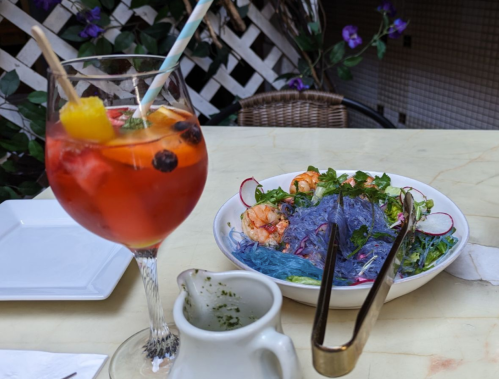 A colorful drink with fruit and a plate of shrimp on blue noodles, garnished with herbs and radishes on a table.