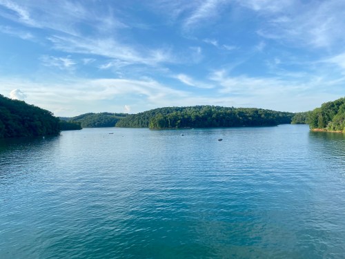 A serene lake surrounded by lush green hills under a clear blue sky. Gentle ripples reflect the landscape.