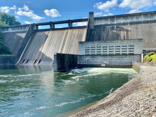 A large concrete dam with water flowing over it, surrounded by green hills and a blue sky with clouds.