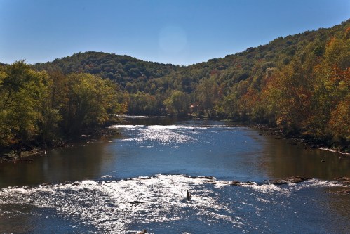 A serene river flows through a lush landscape, reflecting sunlight with hills and trees in the background.
