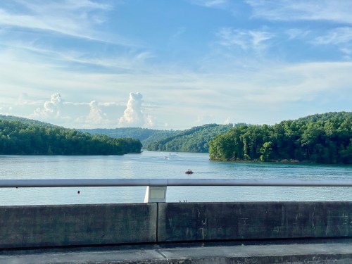 A serene lake surrounded by green hills under a blue sky with fluffy clouds, viewed from a bridge.