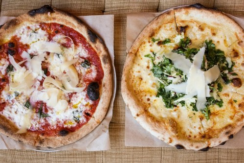 Two pizzas on parchment paper: one with red sauce and cheese, the other with white sauce and greens, both topped with shaved cheese.