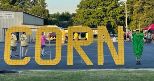A large "CORN" sign made of yellow corn, with a person in a green costume standing next to it at an outdoor event.