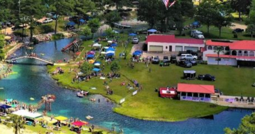 Aerial view of a lively outdoor event by a river, with people, tents, and activities on the grassy area.