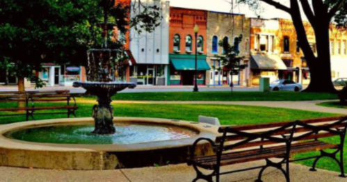 A peaceful park scene featuring a fountain, benches, and a view of quaint storefronts in the background.