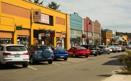 Colorful storefronts line a street with parked cars, under a partly cloudy sky.