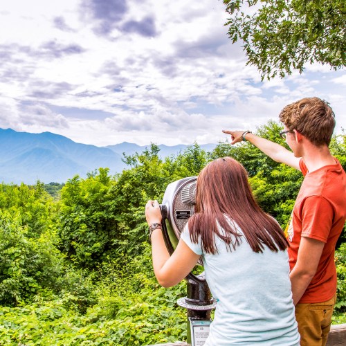 A couple stands at a viewpoint, using a telescope to look at mountains and clouds in the distance.