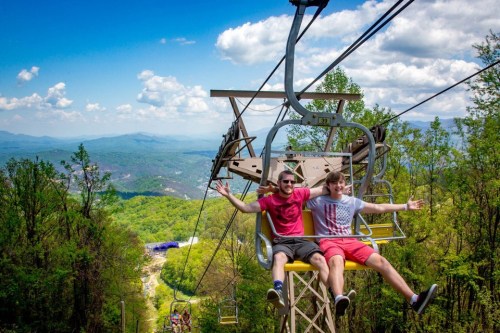 Two friends on a chairlift, smiling and enjoying the scenic mountain view on a sunny day.