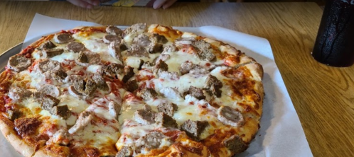 A large pizza topped with cheese, meatballs, and sausage, served on a wooden table with a drink in the background.