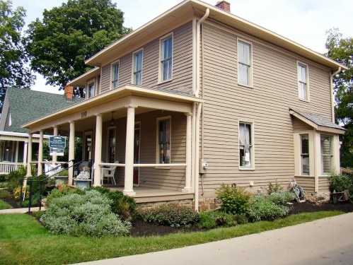 A two-story beige house with a porch, surrounded by greenery and a well-maintained lawn.