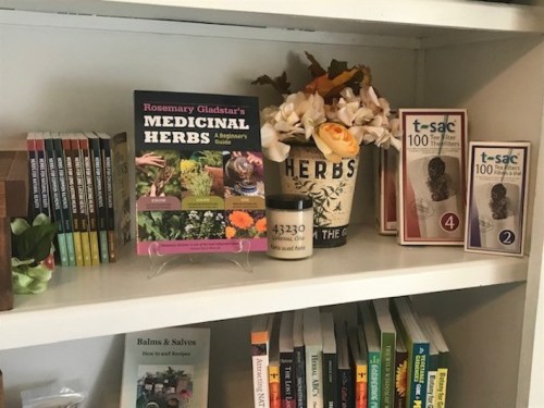 A shelf displaying books on herbs, a candle, and decorative items, including a pot labeled "HERBS."