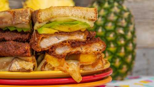 A stack of crispy chicken sandwiches with pineapple and avocado, served on colorful plates with a pineapple in the background.