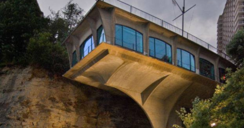 A modern concrete building with large windows, perched on a cliff, surrounded by trees and urban scenery.
