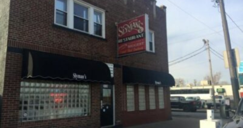 Exterior of a brick building with a sign for "Syd's the Sandwich" and black awnings, located on a city street.