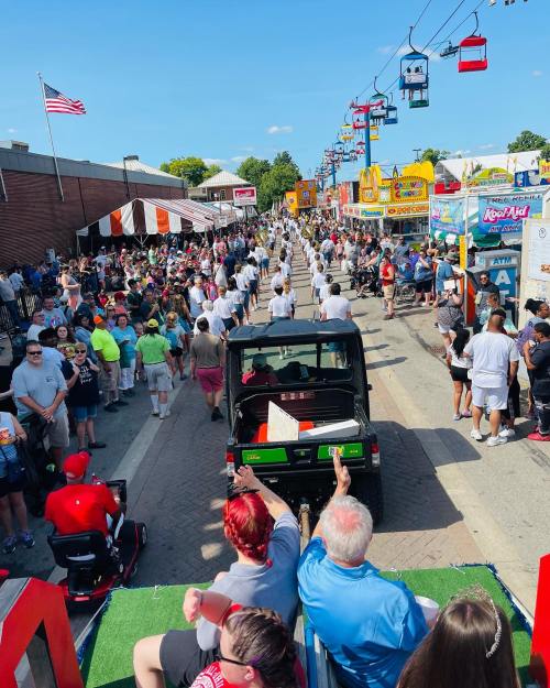 A lively parade scene with crowds, food stalls, and participants walking down a street under a clear blue sky.