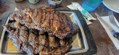 A platter of grilled, seasoned steak with grill marks, served with a side of sauce and a drink.