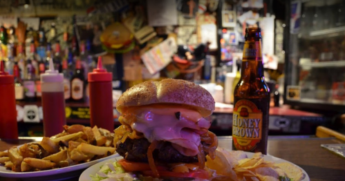 A large burger with toppings, served with fries and a bottle of Honey Brown beer in a lively bar setting.