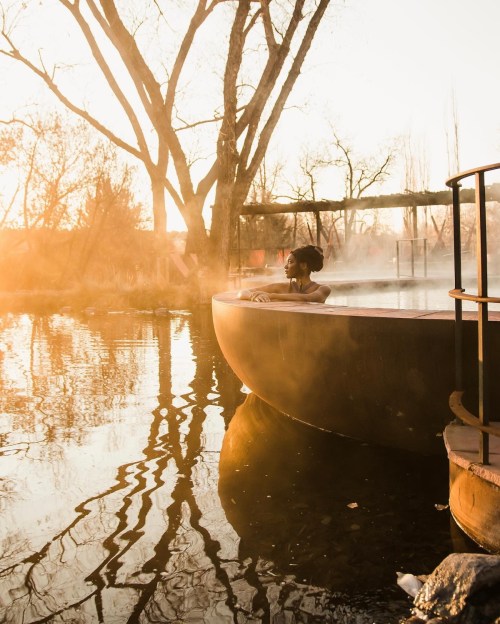 A person relaxes in a steaming hot spring at sunset, surrounded by trees and calm water reflections.