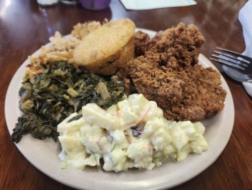 A plate of Southern comfort food featuring fried chicken, cornbread, collard greens, and potato salad.
