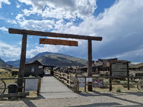 Entrance to Old Trail Town with wooden sign, rustic buildings, and mountains under a partly cloudy sky.