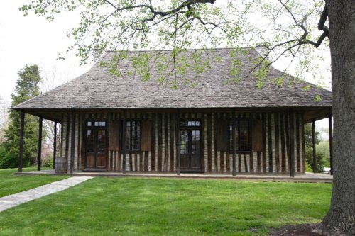 A rustic wooden building with a sloped roof, surrounded by green grass and trees on a sunny day.