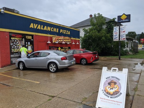 A red car parked outside Avalanche Pizza, with a sign advertising "Holy Shitake! It's Super Mushroom" in the foreground.