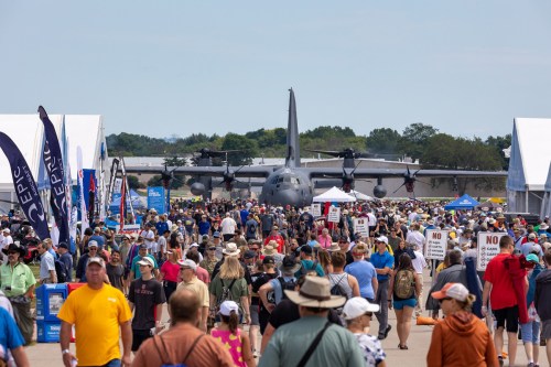 A large crowd gathers at an airshow, with a military aircraft in the background and various tents and banners around.