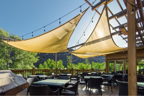 A sunny outdoor patio with shaded seating, string lights, and views of green trees and mountains.