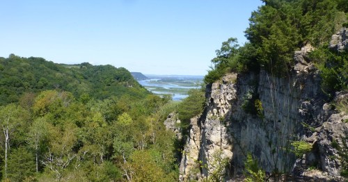A scenic view of a river valley surrounded by lush greenery and rocky cliffs under a clear blue sky.