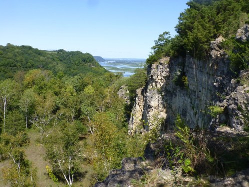 A scenic view of a rocky cliff surrounded by lush greenery and a river in the distance under a clear blue sky.