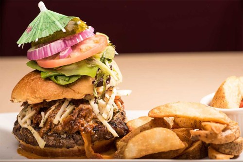 A towering burger topped with vegetables and an umbrella, served with crispy potato wedges on the side.