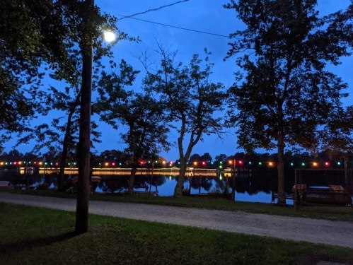 A serene lakeside scene at dusk, with colorful lights strung along trees reflecting on the water.