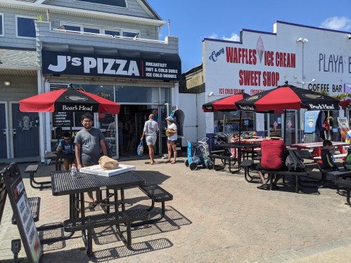 A busy boardwalk scene featuring J's Pizza, outdoor seating, and people enjoying food and drinks on a sunny day.