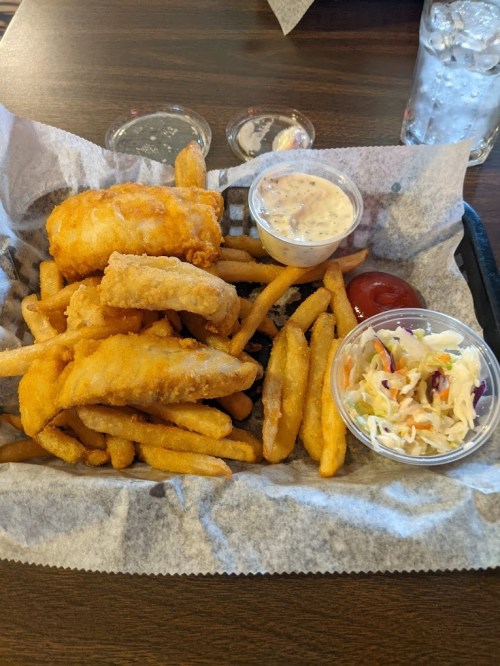A basket of golden fried fish fillets with French fries, coleslaw, and dipping sauces on a wooden table.