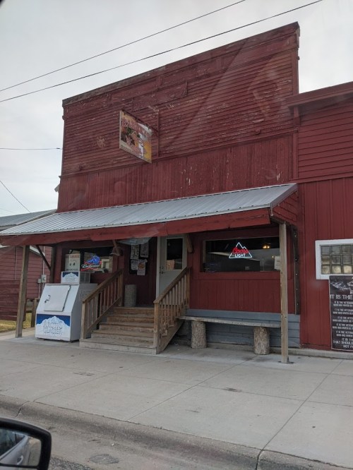 A rustic red building with a wooden porch, featuring a sign and a small entrance, set against a cloudy sky.