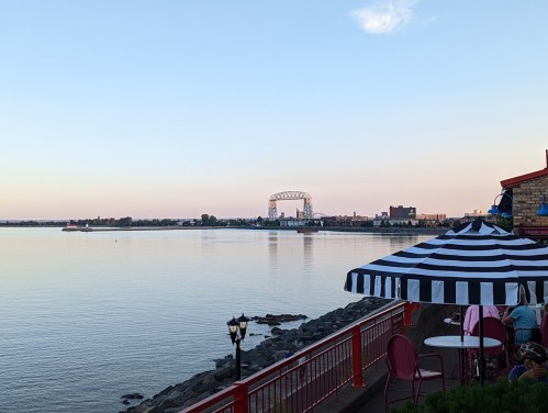 A calm waterfront scene at dusk, featuring a bridge, buildings, and a striped umbrella on the shore.