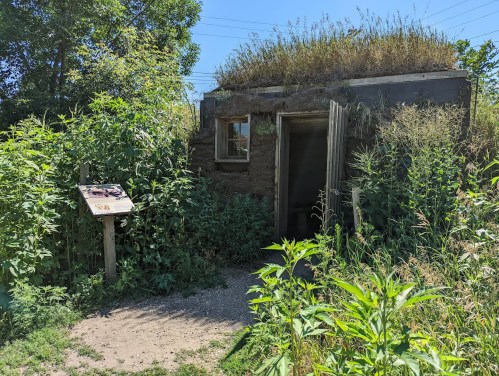 A small, earthy structure with a grass-covered roof, surrounded by tall green plants and a sign nearby.