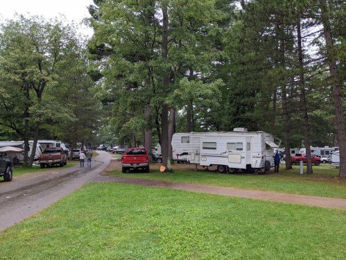 A campground scene with RVs and cars among trees, featuring a gravel path and grassy areas.