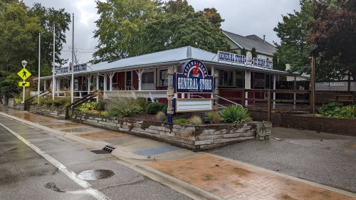 A quaint general store with a blue and white sign, surrounded by greenery on a rainy day.