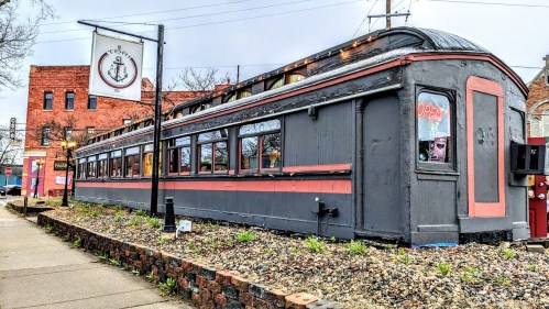 A vintage train car restaurant with a sign reading "Open," surrounded by gravel and buildings in the background.