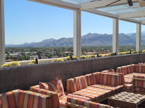 Rooftop lounge with colorful seating, overlooking mountains and palm trees under a clear blue sky.