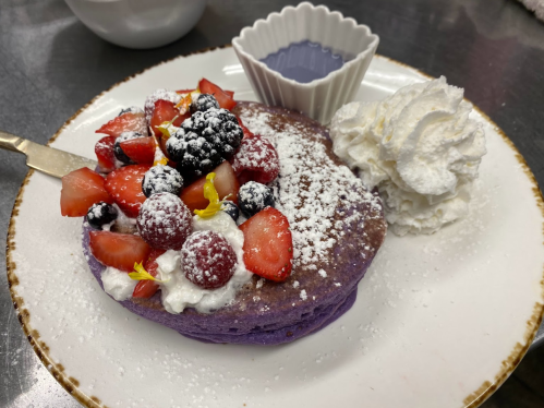 A plate of purple pancakes topped with mixed berries, whipped cream, and a small dish of syrup.