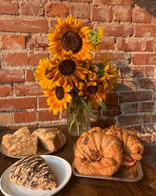 A vase of sunflowers beside a plate of pastries, including croissants and scones, against a brick wall backdrop.