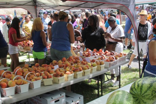 A bustling farmers market with people shopping for peaches and other produce under a tent.