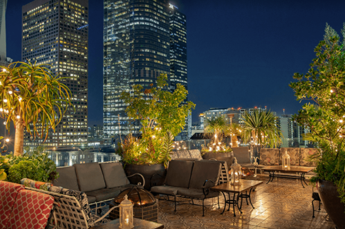 Rooftop lounge at night with cozy seating, greenery, and city skyline illuminated by lights.