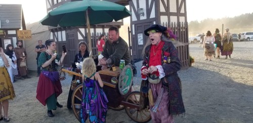 A lively scene at a fair with people in costumes, a cart selling goods, and a colorful character in a hat.
