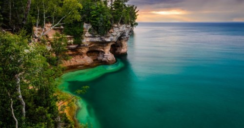 Pink-brown sandstone cliffs with base caves rise from green water under a sunset sky and dense forest.