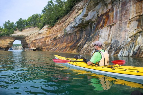A kayaker paddles near colorful cliffs and a natural rock arch, surrounded by calm water and lush greenery.