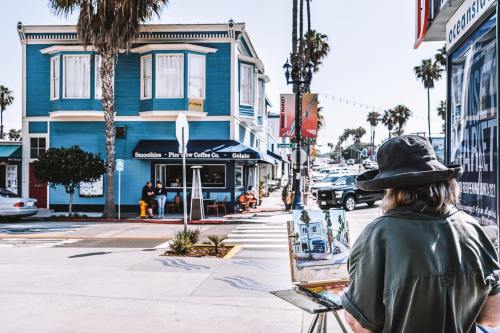 A painter captures a colorful street scene with a blue building and palm trees in a coastal town.